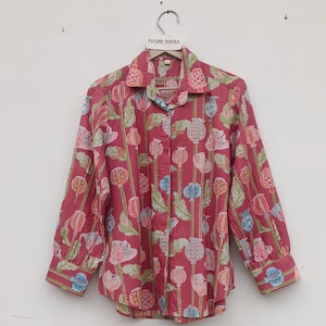 Puede incluir: Una camisa de manga larga con botones en un color rosa intenso, con un estampado botánico repetitivo de cápsulas de amapola y hojas en tonos azules, naranjas y verdes. La camisa tiene cuello y puños estándar, y cuelga de una percha con la etiqueta FUTURE TEXTILE.