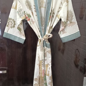 May include: A white cotton robe with a green and white striped trim. The robe has a floral print with green leaves and pink flowers. The robe is tied at the waist with a sash.
