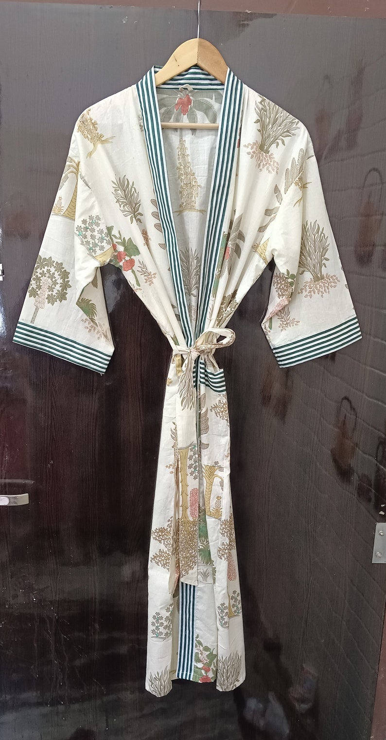 May include: A white cotton robe with a green and white striped trim. The robe has a floral print with green leaves and pink flowers. The robe is tied at the waist with a sash.