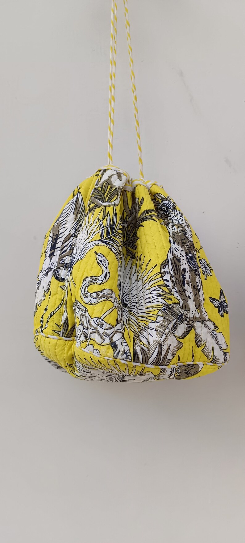May include: A yellow drawstring bag with a white and black floral print featuring snakes, butterflies, and plants. The bag has a white drawstring and is made of quilted fabric.