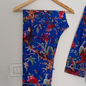 May include: Blue pajama set with a floral and bird print. The set includes pants and a long-sleeved top, both featuring a vibrant design of red flowers, green leaves, and birds on a blue background. White trim outlines the edges of the pajamas.