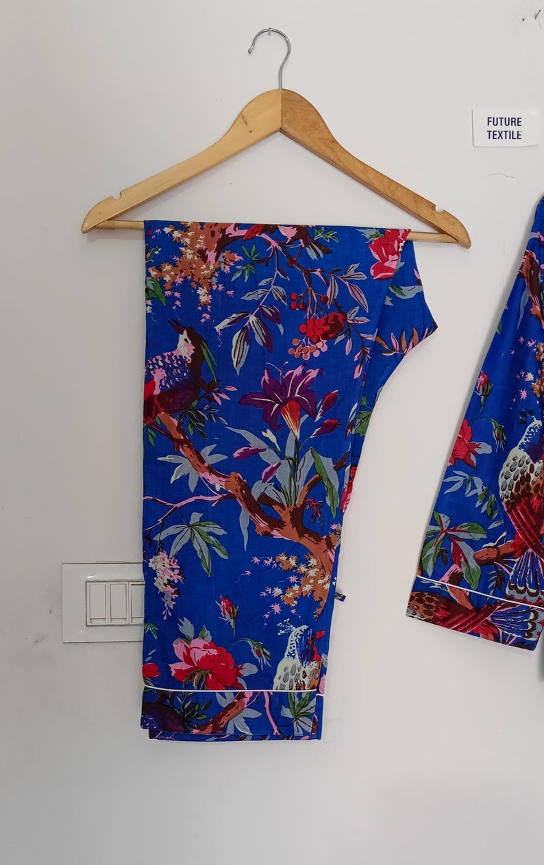 May include: Blue pajama set with a floral and bird print. The set includes pants and a long-sleeved top, both featuring a vibrant design of red flowers, green leaves, and birds on a blue background. White trim outlines the edges of the pajamas.