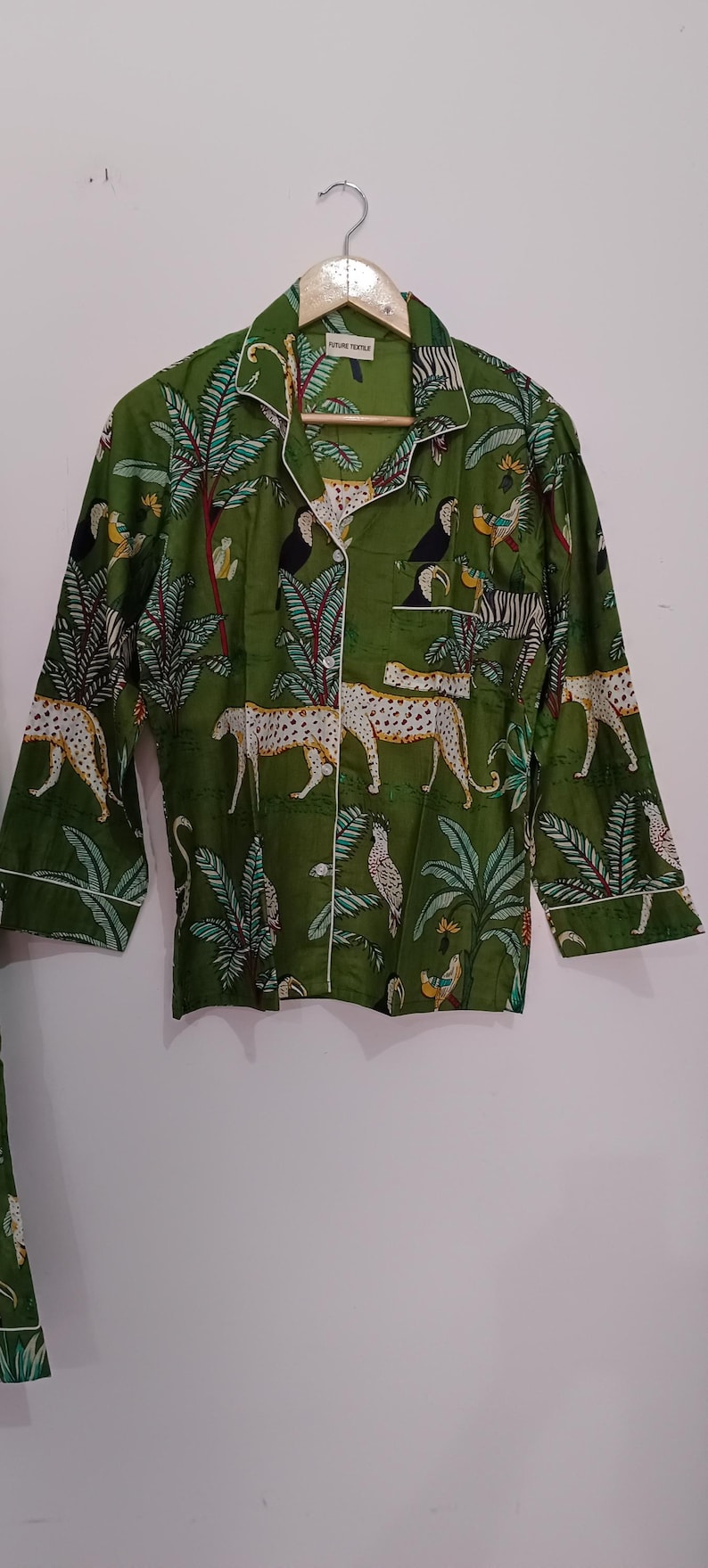 以下が含まれることがあります： 白い縁取りのある緑色のボタンダウンシャツ。シャツには、ヒョウ、オニオオハシ、ヤシの木が描かれたジャングル柄がプリントされています。シャツのラベルには「Future Textile」と書いてあります。