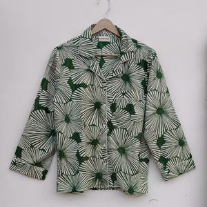 K&ouml;nnte beinhalten: Ein gr&uuml;n-wei&szlig;es Hemd mit Kn&ouml;pfen und einem floralen Muster. Das Hemd hat einen Kragen und lange &Auml;rmel. Das Hemd h&auml;ngt an einem Holzb&uuml;gel.