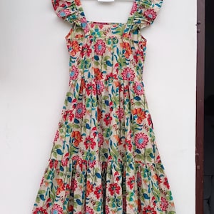 May include: A beige maxi dress with a floral print featuring red, orange, blue, and green flowers. The dress has a square neckline, ruffled straps, and a tiered skirt. The dress is hanging on a wooden hanger with a tag that reads "Future Textile".