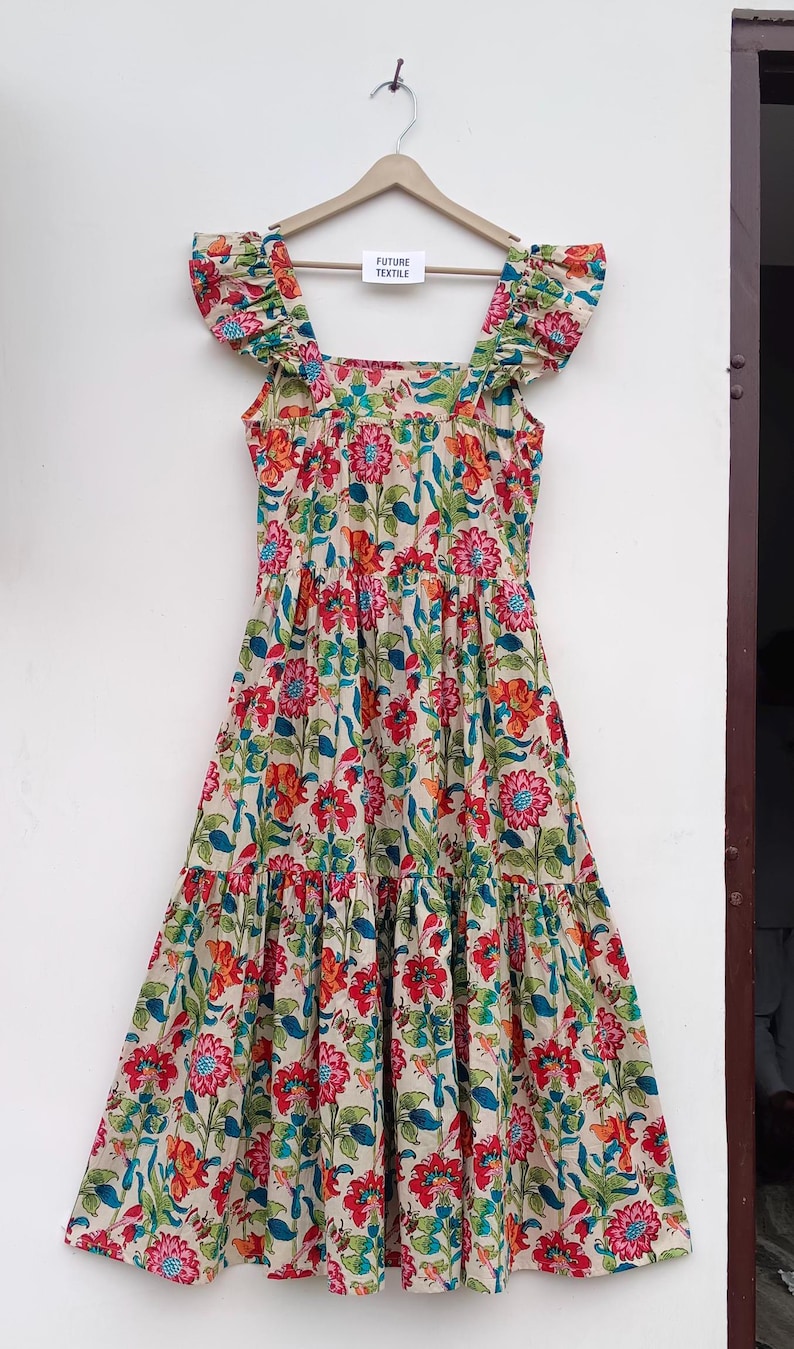 May include: A beige maxi dress with a floral print featuring red, orange, blue, and green flowers. The dress has a square neckline, ruffled straps, and a tiered skirt. The dress is hanging on a wooden hanger with a tag that reads "Future Textile".