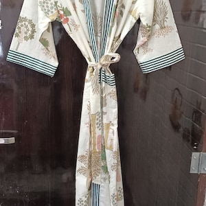 May include: A white cotton robe with a green and white striped trim. The robe features a floral print in a variety of colours, including pink, yellow, and green. The robe is tied at the waist with a matching sash.