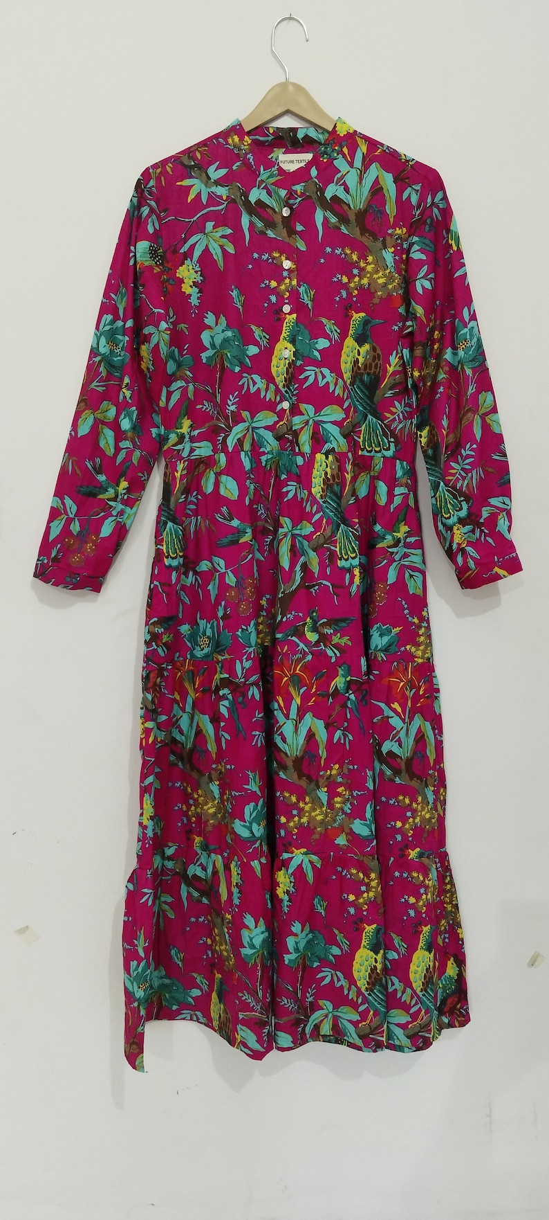 May include: A pink long-sleeved dress with a floral print featuring green leaves, yellow flowers, and colorful birds. The dress has a button-up front and a ruffled hem.