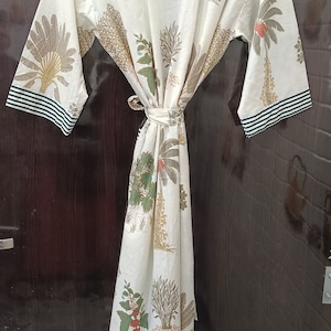 May include: A white cotton robe with a green and white striped trim. The robe features a floral print of palm trees and other tropical plants.