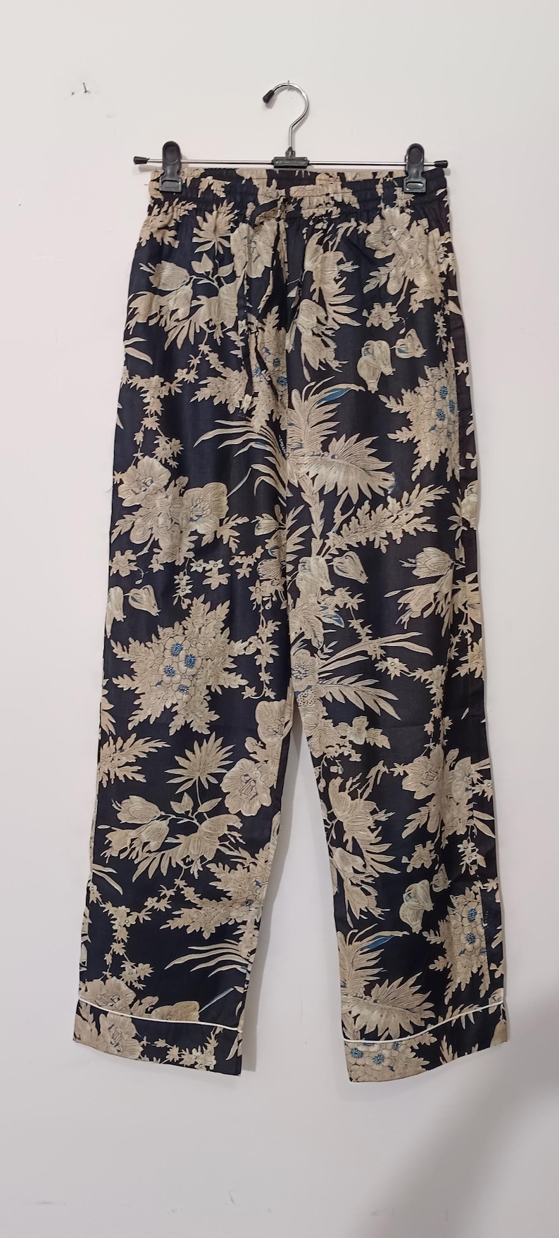 May include: Black pants with a floral print in beige and blue. The pants have a drawstring waist and a white trim.