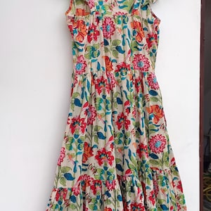 May include: A beige cotton dress with a floral print in shades of red, pink, blue, and green. The dress has a square neckline, ruffled straps, and a tiered skirt.
