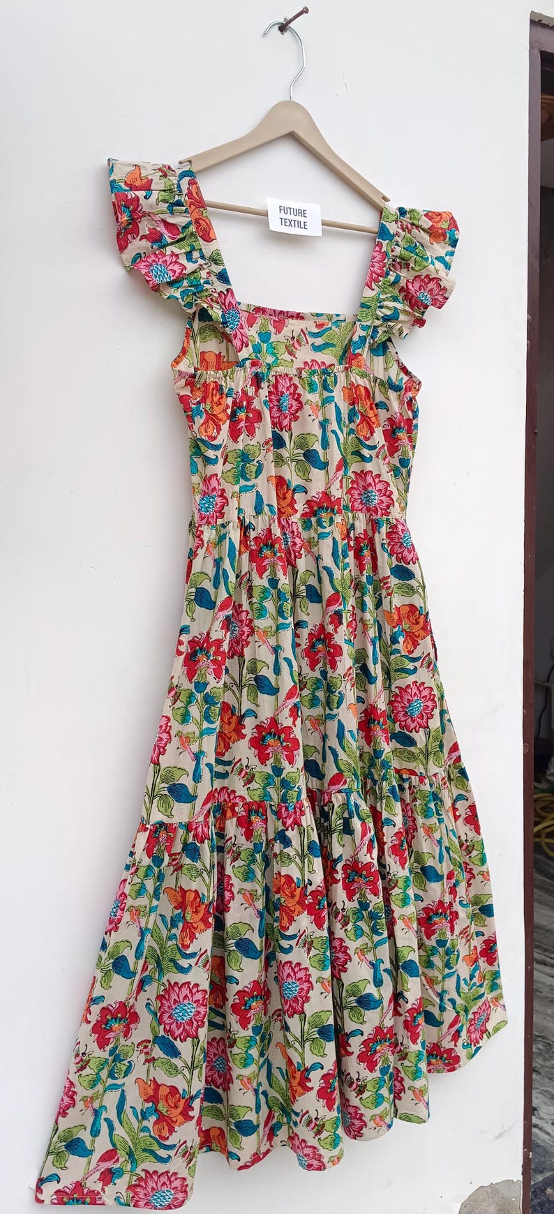 May include: A beige cotton dress with a floral print in shades of red, pink, blue, and green. The dress has a square neckline, ruffled straps, and a tiered skirt.