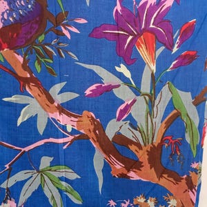 May include: Blue fabric with a vibrant floral and bird pattern. The design features purple lilies, pink and red flowers, and a bird with purple, brown, and blue feathers. The branches are brown, and the leaves are green and gray.