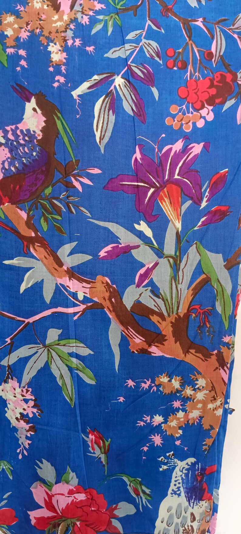 May include: Blue fabric with a vibrant floral and bird pattern. The design features purple lilies, pink and red flowers, and a bird with purple, brown, and blue feathers. The branches are brown, and the leaves are green and gray.