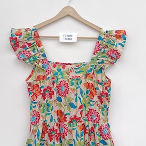 May include: A white cotton dress with a floral print in shades of red, pink, blue, and green. The dress has a square neckline, ruffled straps, and a tiered skirt. The dress is hanging on a wooden hanger with a tag that reads "Future Textile".