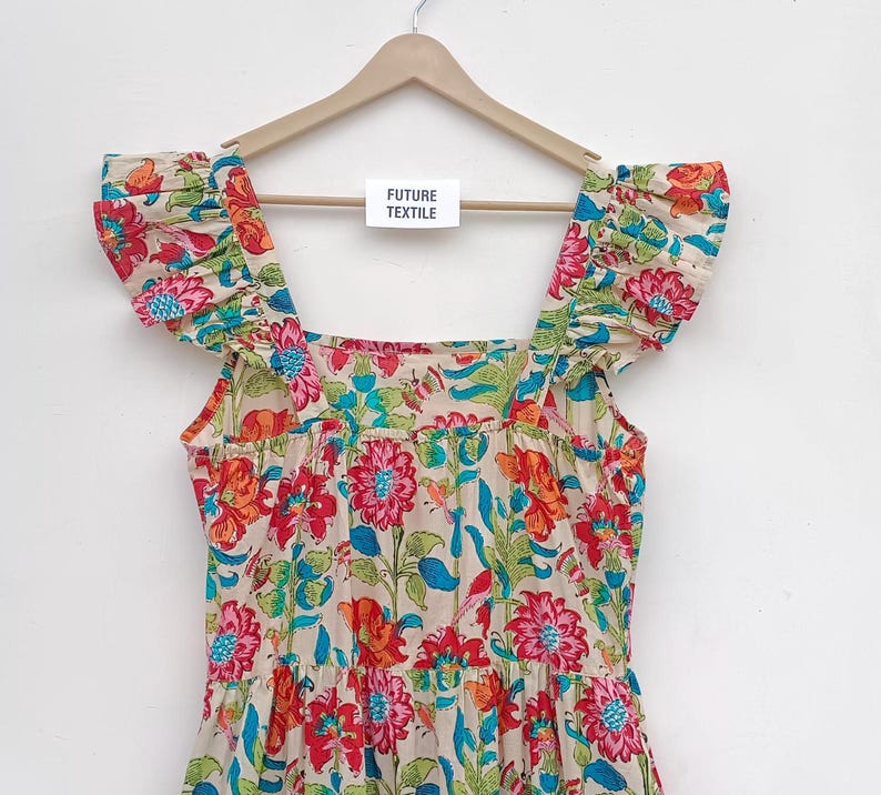 May include: A white cotton dress with a floral print in shades of red, pink, blue, and green. The dress has a square neckline, ruffled straps, and a tiered skirt. The dress is hanging on a wooden hanger with a tag that reads "Future Textile".