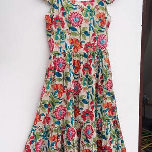 May include: A tiered floral print dress with a square neckline and ruffled cap sleeves. The dress is made of a lightweight fabric and has a relaxed fit. The dress is printed with a colorful floral pattern on a cream background. The dress has a label that says "Future Textile".