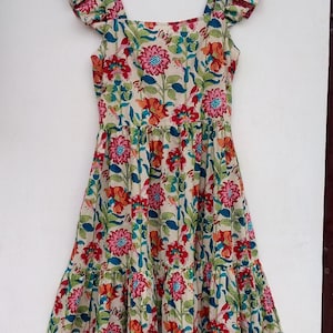 May include: A tiered floral print dress with a square neckline and ruffled cap sleeves. The dress is cream colored with a multi-colored floral pattern. The tag on the dress reads "Future Textile".