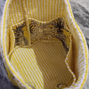 May include: Yellow and white striped fabric tote bag with a floral print lining and multiple pockets. The bag has a drawstring closure.