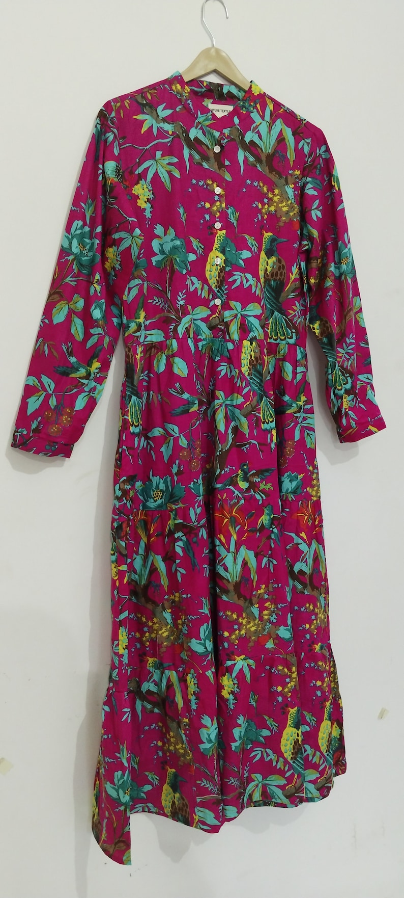 May include: A pink long-sleeved dress with a floral print featuring green leaves and yellow birds. The dress has a button-up front and a gathered skirt. The label on the dress reads "Nature Trails".