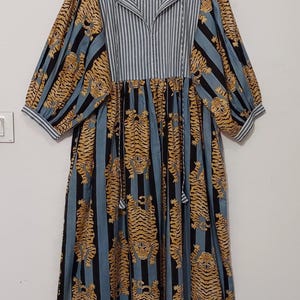May include: A blue and black striped dress with a tiger print. The dress has a V-neck and 3/4 sleeves. The dress is made of cotton and has a relaxed fit. The dress is hanging on a wooden hanger. The label on the dress says "Future People".