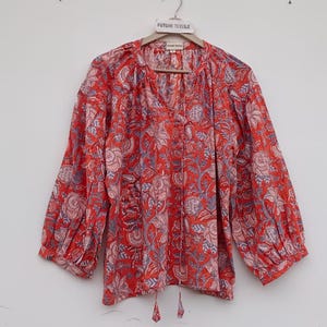 Peut inclure: Un chemisier rouge avec un motif floral dans les tons de rose et de bleu est suspendu à un cintre en bois. Le haut à manches longues a un col en V et un cordon de serrage en bas. Le tissu semble léger et aéré.