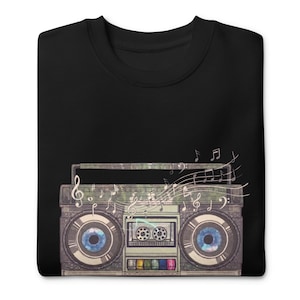 May include: A black crew neck t-shirt featuring a graphic of a vintage boombox. The boombox speakers are depicted as large blue human eyes, and musical notes and staff lines swirl out from the top of the device in a light, artistic design.