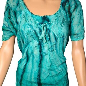 May include: A short-sleeved top in a vibrant turquoise and dark teal tie-dye pattern. The garment features a scoop neckline with a delicate crocheted trim, a central lace-up detail with two small tassels, and intricate tonal embroidery accents.