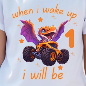 May include: White t-shirt featuring a cartoon dragon in an orange monster truck. The dragon has purple wings and an open mouth. The text "when i wake up i will be 1" is in orange.