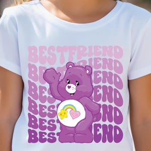 May include: White t-shirt with a purple Care Bear wearing a rainbow and a heart on its belly. The words "BEST FRIEND" are repeated in pink on the shirt.