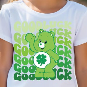 May include: White t-shirt with a green Care Bear wearing a four-leaf clover and waving. The shirt has the words "Good Luck" repeated in a green gradient pattern.