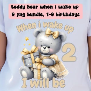 May include: A grey teddy bear wearing a gold dress and holding a gift box. The bear has a gold bow in its hair and the text "When I wake up I will be 2" is written on the image.