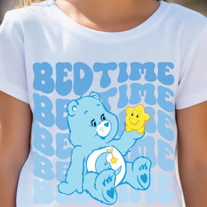May include: A white t-shirt with a blue Care Bear holding a yellow star. The shirt has the text "BEDTIME" repeated in a blue retro font.