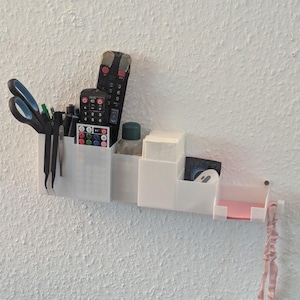 Può includere: Un organizer bianco da parete con più scomparti. Contiene vari oggetti, tra cui forbici, penne, telecomandi, una bottiglietta e fazzoletti. Un oggetto decorativo rosa e grigio è appeso al lato.