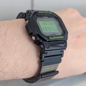 Könnte beinhalten: Schwarze digitale G-Shock-Uhr mit grüner Digitalanzeige. Die Uhr hat ein schwarzes Armband mit einem grün-beigen Stoffband. Auf dem Zifferblatt sind die Wörter "PROTECTION", "CASIO", "TOUGH SOLAR" und "G-SHOCK" aufgedruckt.