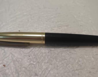 May include: A gold-colored pen with a black barrel. The pen has a gold-colored clip and a black tip. The pen is lying on a white surface. The pen is a writing instrument.