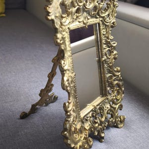 Peut inclure: Miroir doré orné avec un cadre en métal. Le miroir rectangulaire est soutenu par un support décoratif. Le cadre présente un design vintage élaboré avec un chérubin au sommet.