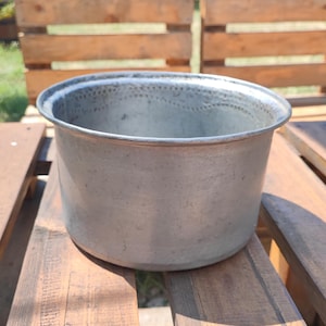 Op de afbeelding: Een ronde, zilverkleurige metalen pot met een brede rand en een licht getextureerd oppervlak. De pot heeft een eenvoudig, functioneel ontwerp, geschikt om in te koken of op te bergen. De pot staat op een houten oppervlak.