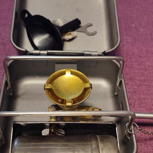 May include: A silver camping stove with a brass burner and a silver fuel canister. The stove is in a gray metal case with a black lid. The case also contains a black metal funnel and a wrench.
