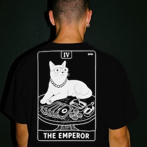 Puede incluir: Una camiseta negra con un estampado gráfico blanco de un gato con gafas y una cadena, sentado a una mesa con dinero y latas. El diseño se asemeja a una carta del tarot etiquetada como IV y THE EMPEROR, con el texto Sable visible en la esquina.