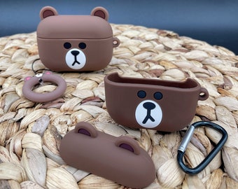 May include: Brown silicone case for AirPods with a cartoon bear face design. The case has a detachable carabiner and a ring for attaching to a keychain.