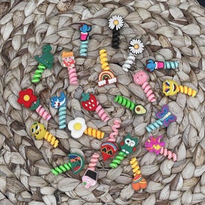 May include: A collection of colorful cable organizers with various cute designs, including a cactus, a dinosaur, a strawberry, a rainbow, a flower, an avocado, a butterfly, an owl, and a heart. The organizers are made of flexible, coiled plastic and are designed to keep cords and cables tidy.