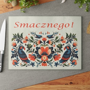 May include: A white cutting board with a colorful floral design featuring red, blue, and green flowers, leaves, and birds. The text "Smacznego!" is printed in red at the top of the board.
