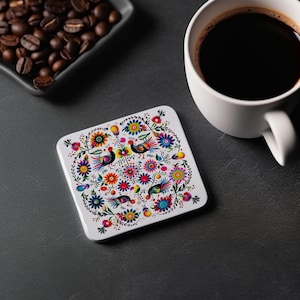 May include: A white square coaster with a colorful floral and bird design. The coaster features a repeating pattern of flowers, leaves, and birds in shades of red, yellow, blue, green, and pink.
