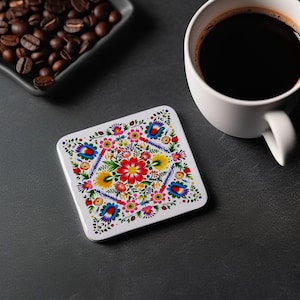 Puede incluir: Un posavasos cuadrado blanco con un diseño floral colorido. El posavasos presenta una flor central con pétalos rojos y centro amarillo rodeado de flores más pequeñas en tonos de rosa, azul, amarillo y verde.