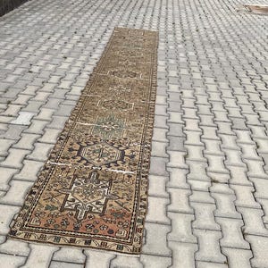 Peut inclure: Un long tapis rectangulaire vintage avec un motif g&eacute;om&eacute;trique d&eacute;taill&eacute; dans des tons de marron, beige et des touches de vert et de bleu. Le tapis est pos&eacute; sur une surface en briques grises, mettant en valeur son design complexe et son aspect vieilli.