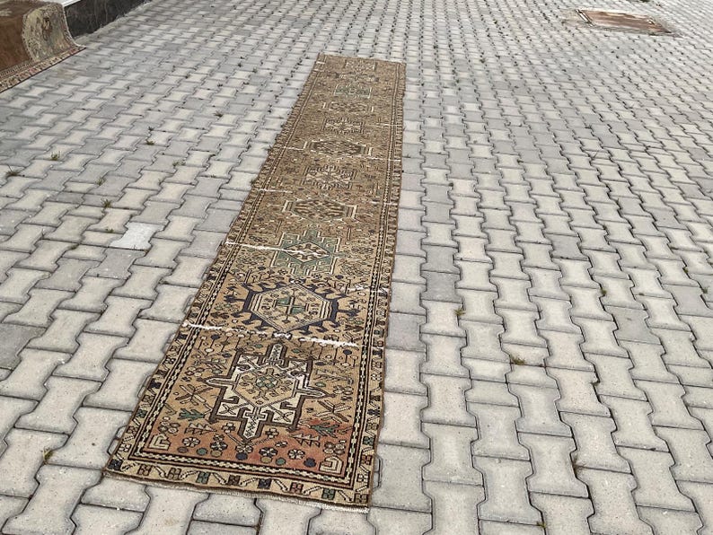 Peut inclure: Un long tapis rectangulaire vintage avec un motif g&eacute;om&eacute;trique d&eacute;taill&eacute; dans des tons de marron, beige et des touches de vert et de bleu. Le tapis est pos&eacute; sur une surface en briques grises, mettant en valeur son design complexe et son aspect vieilli.