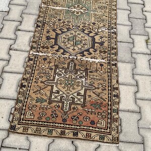 Peut inclure: Un tapis rectangulaire de style vintage avec un motif g&eacute;om&eacute;trique d&eacute;taill&eacute;. Le tapis pr&eacute;sente un motif central dans des tons beige, marron et vert, avec une bordure bleu fonc&eacute;. Le tapis est pos&eacute; sur une surface gris clair.
