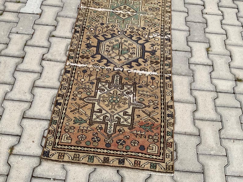 Peut inclure: Un tapis rectangulaire de style vintage avec un motif g&eacute;om&eacute;trique d&eacute;taill&eacute;. Le tapis pr&eacute;sente un motif central dans des tons beige, marron et vert, avec une bordure bleu fonc&eacute;. Le tapis est pos&eacute; sur une surface gris clair.