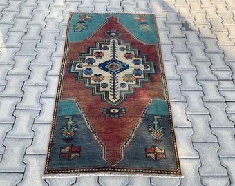 Könnte beinhalten: Rechteckiger Teppich mit einem zentralen Rautenmuster in Rot-, Blau- und Cremetönen. Der Teppich weist geometrische Designs und einen türkisfarbenen Rand auf. Der Teppich liegt auf einer grauen Oberfläche.